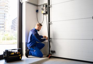 A technician in a blue uniform kneeling next to a white sectional garage door, adjusting the opener system with a toolbox nearby in a Mahtomedi garage.