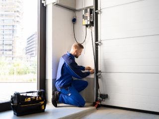 Technician Performing Garage Door Maintenance – Mahtomedi, MN A technician in a blue uniform kneeling next to a white sectional garage door, adjusting the opener system with a toolbox nearby in a Mahtomedi garage.