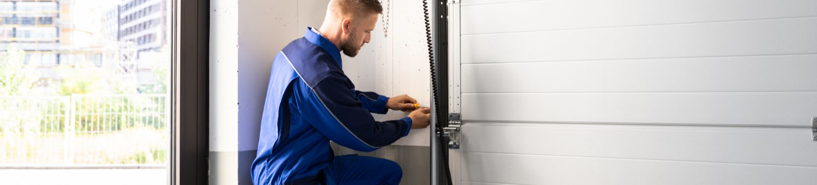 A technician in a blue uniform kneeling next to a white sectional garage door, adjusting the opener system with a toolbox nearby in a Mahtomedi garage.