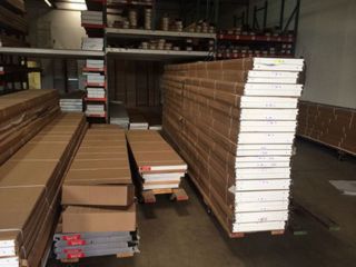 Garage Door Panels and Parts in Warehouse – Selection Guide Stacks of boxed garage door panels and hardware organized on pallets in a well-lit warehouse, ready for selection and installation.